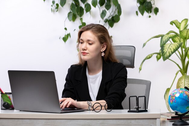 Eine junge schöne Dame der Vorderansicht im weißen Hemd und in der schwarzen Jacke, die mit Dokumenten unter Verwendung ihres Laptops vor Tisch mit hängenden Blättern arbeiten
