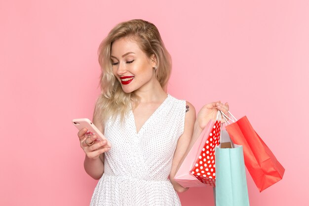 Eine junge schöne Dame der Vorderansicht im weißen Kleid, die Einkaufspakete hält und ein Telefon mit einem Lächeln auf ihrem Gesicht benutzt