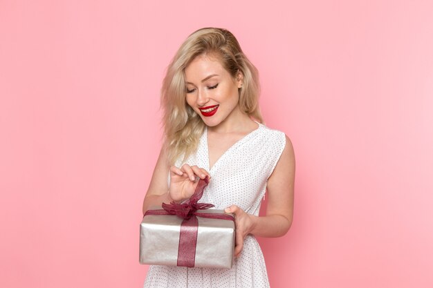 Eine junge schöne Dame der Vorderansicht im weißen Kleid, die Geschenkbox mit Lächeln auf ihrem Gesicht öffnet