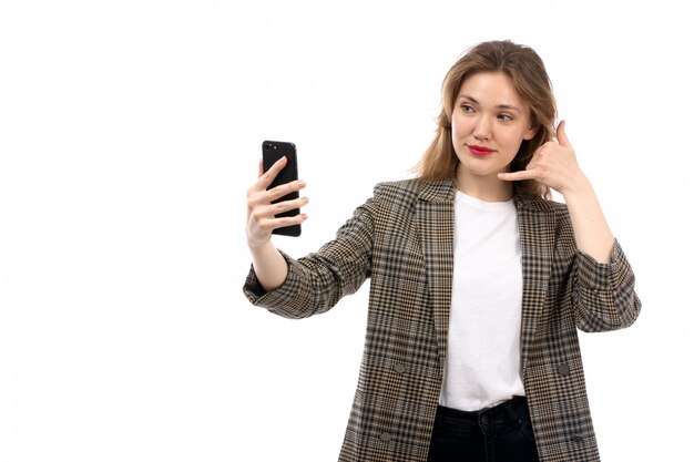 Eine junge schöne Dame der Vorderansicht in den schwarzen Jeans und im Mantel des weißen T-Shirts unter Verwendung des Smartphones auf dem Weiß