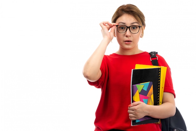 Eine junge schöne Dame der Vorderansicht in der schwarzen Jeans des roten T-Shirts, die verschiedene Hefte hält und Brillen mit Tasche auf dem Weiß feilt