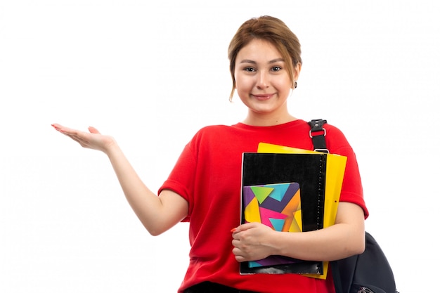 Eine junge schöne dame der vorderansicht in der schwarzen jeans des roten t-shirts, die verschiedene hefte und dateien hält, die mit tasche auf dem weiß lächeln