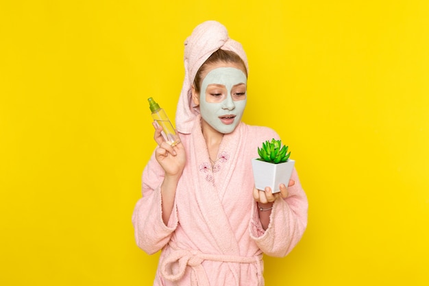 Eine junge schöne Frau der Vorderansicht im rosa Bademantel, der Spray und kleine Pflanze hält