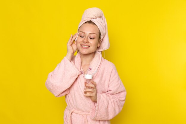 Eine junge schöne Frau der Vorderansicht im rosa Bademantel, die ihr ganzes Gesicht mit Reiniger aufräumt