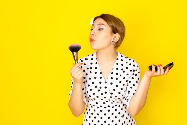 Eine junge schöne Frau der Vorderansicht im Schwarzweiss-Tupfenkleid, das Make-up auf Mode-Mascara-Pinsel der gelben Hintergrundkleidung macht