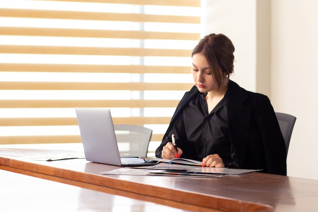 Eine junge schöne geschäftsfrau der vorderansicht in der schwarzen jacke des schwarzen hemdes unter verwendung ihrer silbernen laptop-schreiblesung, die in ihrem büroarbeitsjobgebäude arbeitet