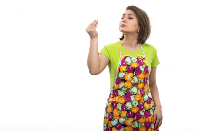 Kostenloses Foto eine junge schöne hausfrau der vorderansicht des bunten umhangs des grünen hemdes, das das leckere zeichen auf der weiblichen küche des weißen hintergrundhauses aufwirft