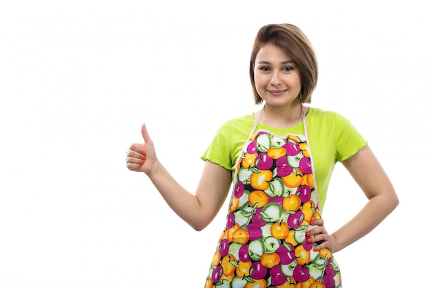 Eine junge schöne Hausfrau der Vorderansicht des bunten Umhangs des grünen Hemdes, das fantastisches Zeichen zeigt, das auf der weiblichen Küche des weißen Hintergrundhauses lächelt