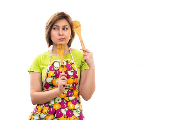 Eine junge schöne Hausfrau der Vorderansicht des bunten Umhangs des grünen Hemdes, der hölzernes Küchengerät denkt, das auf dem weißen Hintergrund Hausreinigungsküche denkt