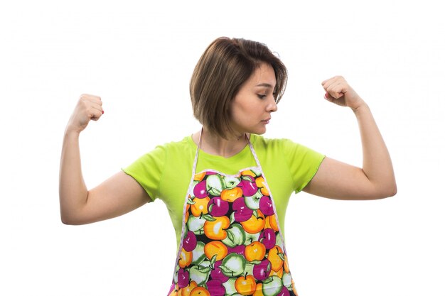 Eine junge schöne Hausfrau der Vorderansicht des bunten Umhangs des grünen Hemdes, der stark auf der weiblichen Küche des weißen Hintergrundhauses biegt