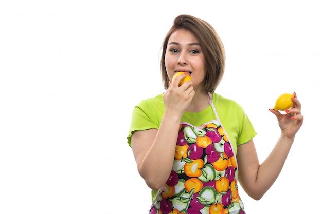 Eine junge schöne Hausfrau der Vorderansicht im bunten Umhang des grünen Hemdes, das Zitronen hält, die auf der weiblichen Küche des weißen Hintergrundhauses versuchen