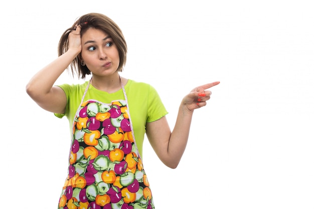 Eine junge schöne Hausfrau der Vorderansicht im bunten Umhang des grünen Hemdes, die überrascht zögerte expressio auf der weiblichen Küche des weißen Hintergrundhauses aufwirft