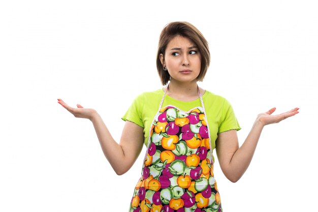 Eine junge schöne Hausfrau der Vorderansicht im bunten Umhang des grünen Hemdes, die überraschten zögernden Ausdruck aufwirft, der glücklich auf der weiblichen Küche des weißen Hintergrundhauses lächelt
