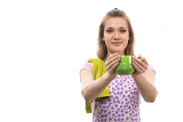 Eine junge schöne Hausfrau der Vorderansicht im bunten Umhang des rosa Hemdes, der die lächelnde Aufstellung der grünen Tasse hält