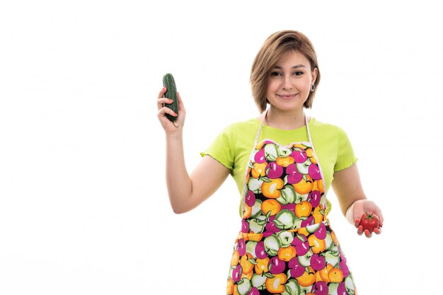 Eine junge schöne Hausfrau der Vorderansicht in buntem Umhang des grünen Hemdes lächelnd, das Gemüse auf der weißen Hintergrundhausreinigungsküche hält