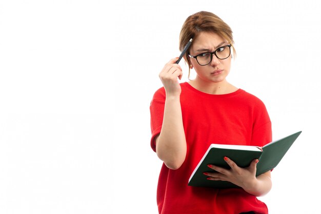 Eine junge Studentin der Vorderansicht im roten T-Shirt, das das Heft hält, das Notizen schreibt, die auf dem Weiß denken