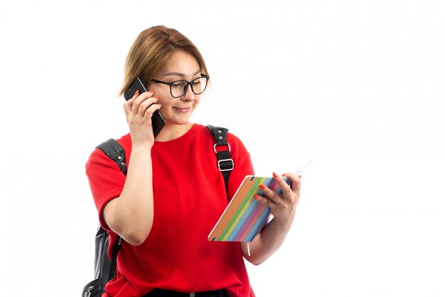 Eine junge Studentin der Vorderansicht im roten T-Shirt, das schwarze Tasche hält, die Heft und schwarzes Smartphone hält, das das Weiß spricht