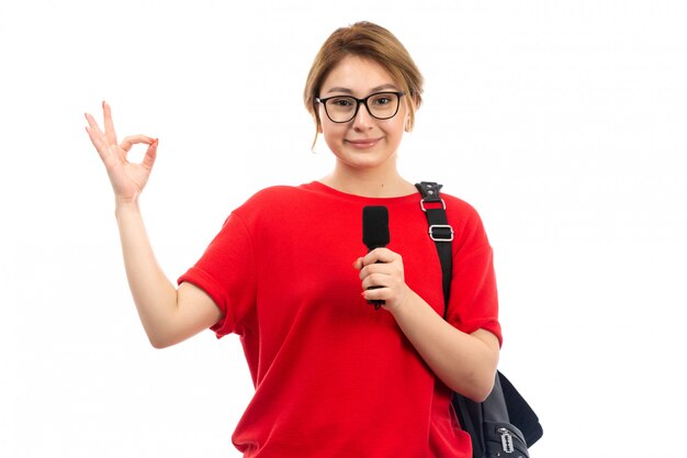Eine junge Studentin der Vorderansicht in der roten T-Shirt tragenden schwarzen Tasche, die das Mikrofon hält, das auf dem Weiß lächelt