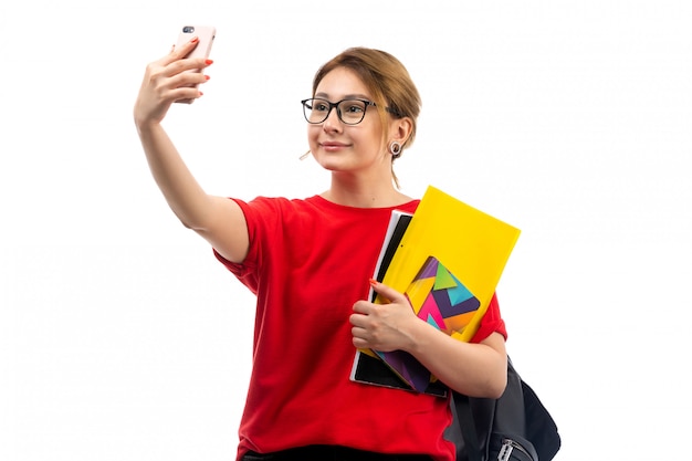 Eine junge Studentin der Vorderansicht in der schwarzen Jeans des roten T-Shirts, die Hefte hält, die ein Selfie auf dem Weiß nehmen