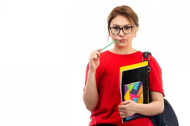 Eine junge Studentin der Vorderansicht in der schwarzen Jeans des roten T-Shirts, die verschiedene Hefte und Dateien hält, die Bleistift denken auf dem Weiß halten