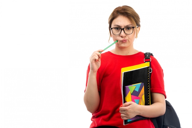 Eine junge Studentin der Vorderansicht in der schwarzen Jeans des roten T-Shirts, die verschiedene Hefte und Dateien hält, die Bleistift denken auf dem Weiß halten