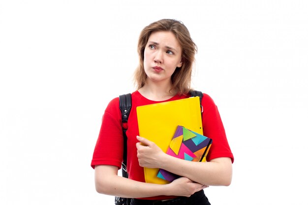 Eine junge Studentin der Vorderansicht in der schwarzen Tasche des roten Hemdes, die Copybook-Dateien hält, die auf dem Weiß denken