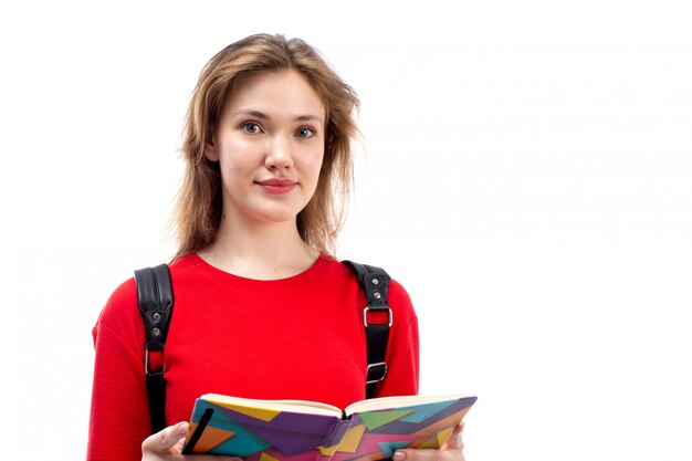 Eine junge Studentin der Vorderansicht in der schwarzen Tasche des roten Hemdes, die Copybook-Dateien hält, die lächelnd auf das Weiß schreiben