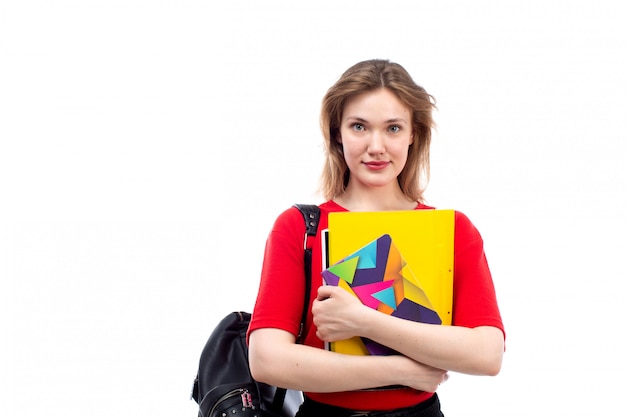 Eine junge Studentin der Vorderansicht in der schwarzen Tasche des roten Hemdes, die die auf dem Weiß lächelnden Hefte hält