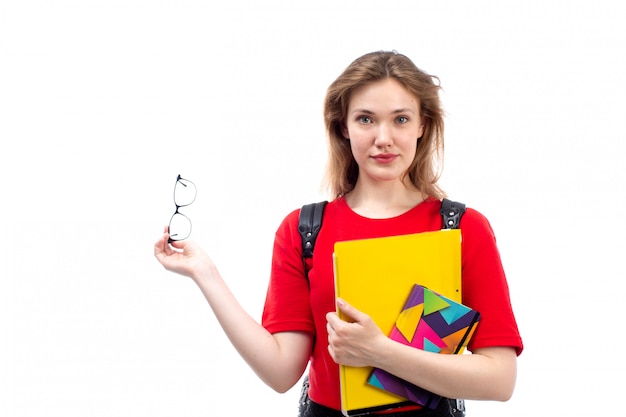 Eine junge Studentin der Vorderansicht in der schwarzen Tasche des roten Hemdes, die die Heftdateien hält, die auf dem Weiß lächeln