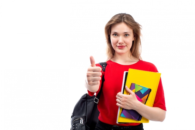 Eine junge Studentin der Vorderansicht in der schwarzen Tasche des roten Hemdes, die Stift und Hefte hält, die auf dem Weiß lächeln