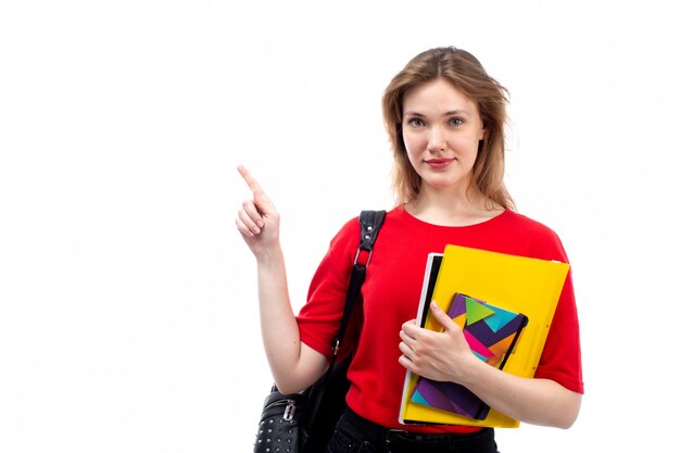 Eine junge Studentin der Vorderansicht in der schwarzen Tasche des roten Hemdes, die Stift und Hefte hält, die lächelnd auf dem Weiß posieren