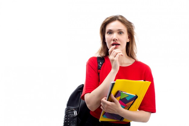 Eine junge Studentin der Vorderansicht in der schwarzen Tasche des roten Hemdes, die Stift und Hefte hält, überrascht auf dem Weiß