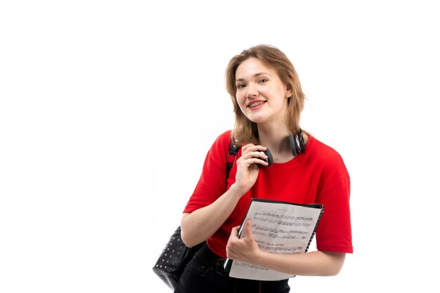 Eine junge Studentin der Vorderansicht in der schwarzen Tasche des roten Hemdes mit den schwarzen Kopfhörern, die Musik hören, die das Copybook auf dem Weiß hält