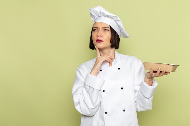 Eine junge weibliche Köchin der Vorderansicht im weißen Kochanzug und im Kappenhaltepaket mit denkendem Ausdruck auf der grünen Wanddamenarbeitsnahrungsmittelküchenfarbe