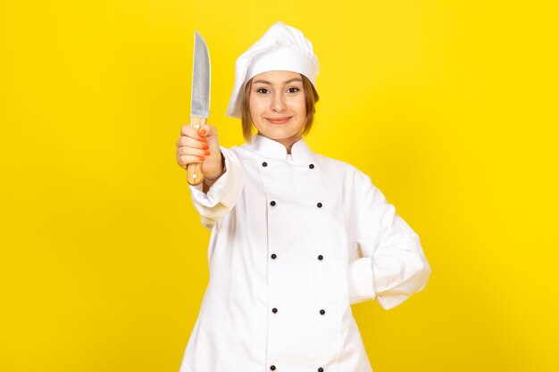 Eine junge weibliche Köchin der Vorderansicht im weißen Kochanzug und im weißen Kappenhaltemesser, das auf dem Gelb lächelt