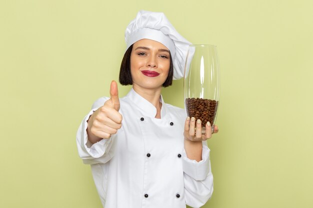 Eine junge weibliche Köchin der Vorderansicht im weißen Kochanzug und in der Kappe, die Glas mit Kaffeesamen auf der grünen Wanddamenarbeitslebensmittelfarbe hält