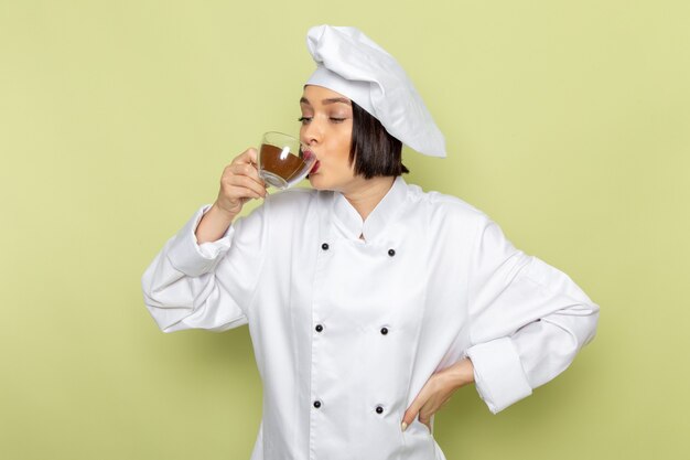 Eine junge weibliche Köchin der Vorderansicht im weißen Kochanzug und in der Kappe, die Tasse des Kaffeepulvers auf der grünen Wand halten und trinken