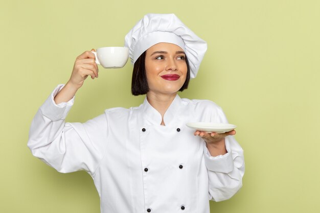 Eine junge weibliche Köchin der Vorderansicht im weißen Kochanzug und in der Kappe, die Tasse Kaffee an der grünen Wand halten