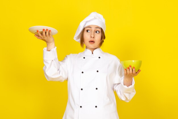Eine junge weibliche Köchin der Vorderansicht im weißen Kochanzug und in der weißen Kappe, die gelbe und rote Platten halten, die auf dem gelben träumen