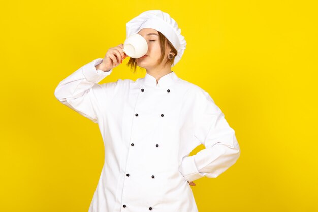 Eine junge weibliche Köchin der Vorderansicht im weißen Kochanzug und in der weißen Kappe, die Kaffee auf dem gelben trinkt