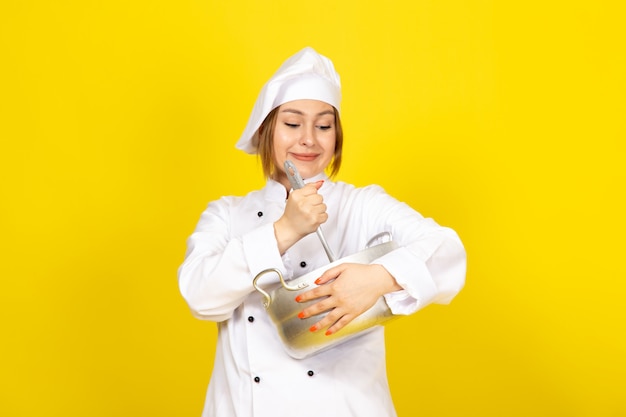 Eine junge weibliche Köchin der Vorderansicht im weißen Kochanzug und in der weißen Kappe, die runde silberne Pfanne hält, die auf dem gelben lächelt