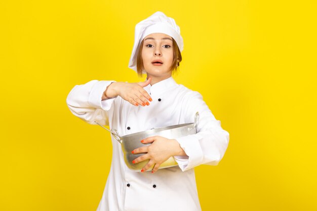 Eine junge weibliche Köchin der Vorderansicht im weißen Kochanzug und in der weißen Kappe, die runde silberne Pfanne hält, die es auf dem gelben mischt