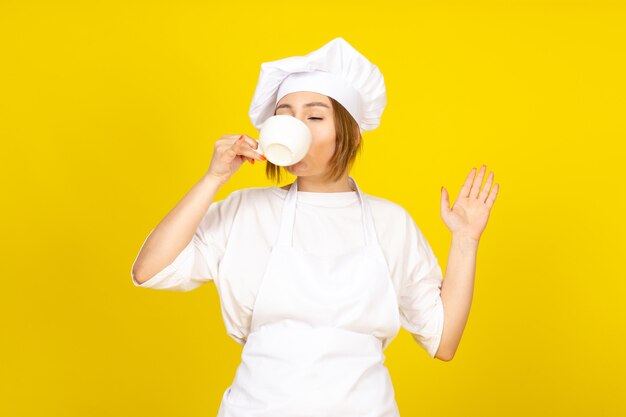 Eine junge weibliche Köchin der Vorderansicht im weißen Kochanzug und in der weißen Kappe, die weißen Tassetrinkkaffee hält, aufgeregt lustig auf dem Gelb