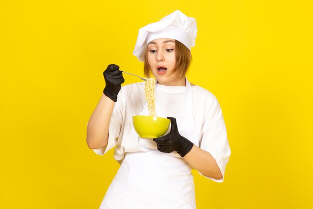 Eine junge weibliche Köchin der Vorderansicht im weißen Kochanzug und in der weißen Kappe in den schwarzen Handschuhen, die grünen Teller halten, der Spaghetti auf dem gelben mischt