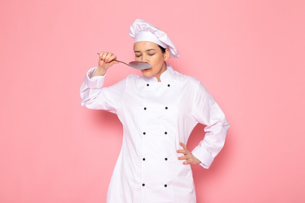 Eine junge weibliche Köchin der Vorderansicht in der weißen Kappe des weißen Kochanzugs, die das schmeckende Essen des großen silbernen Löffels aufwirft