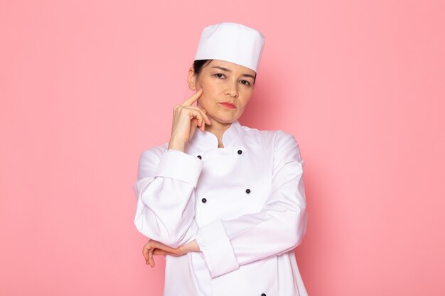 Eine junge weibliche Köchin der Vorderansicht in der weißen Kappe des weißen Kochanzugs, die tief denkenden Ausdruck aufwirft