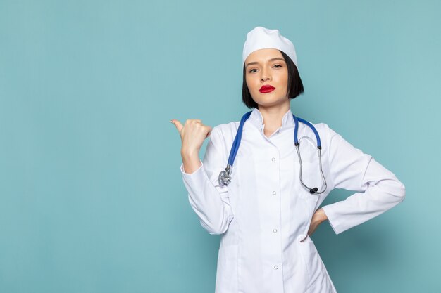 Eine junge weibliche Krankenschwester der Vorderansicht im weißen medizinischen Anzug und im blauen Stethoskop, die auf der blauen Schreibtischmedizin-Krankenhausarztfarbe aufwerfen