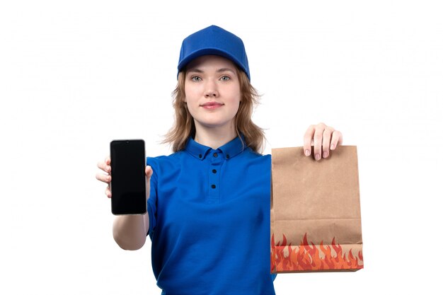 Eine junge weibliche Kurierin der Vorderansicht des weiblichen Lieferservices des Lebensmittellieferservices lächelnd, das Paketlebensmittel und das Smartphone lächelnd auf Weiß hält