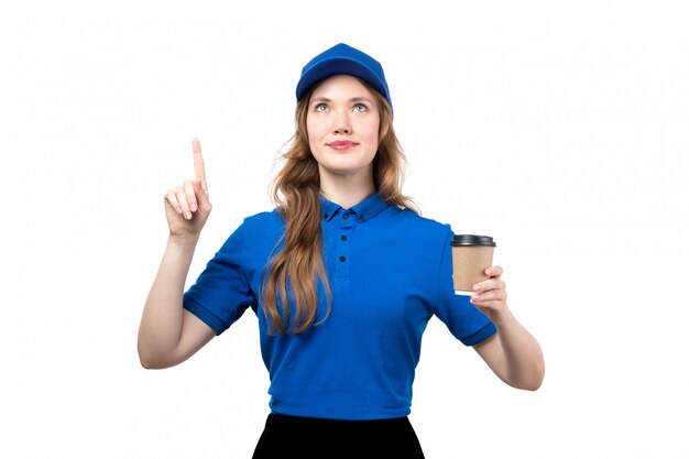 Eine junge weibliche Kurierin der Vorderansicht in der blauen Kappe des blauen Hemdes und der schwarzen Hose lächelnd, die in den Himmel schaut, der Tasse Kaffee auf Weiß hält