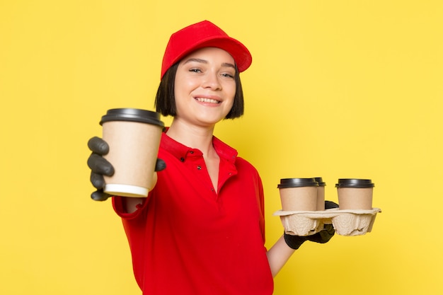 Eine junge weibliche Kurierin der Vorderansicht in schwarzen schwarzen Handschuhen der Uniform und der roten Kappe, die Kaffeetassen hält